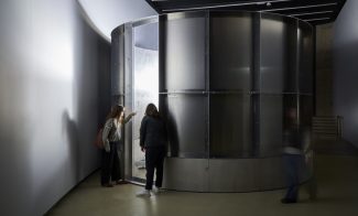 Two people stand closely together, peering into a slightly open door of a large, circular metal structure inside a dimly lit, modern gallery space. Another blurred figure walks by on the right.