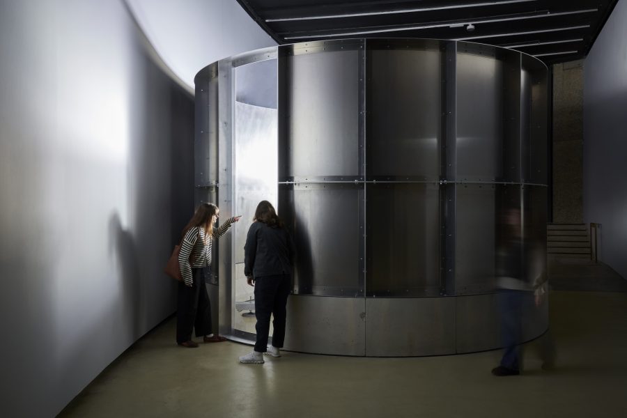 Two people stand closely together, peering into a slightly open door of a large, circular metal structure inside a dimly lit, modern gallery space. Another blurred figure walks by on the right.