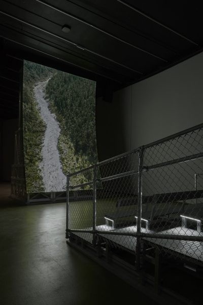 A large photograph of a forested mountain with a rocky stream is displayed vertically in a dimly lit room, behind a metal chain-link barrier and an elevated walkway with spotlights.