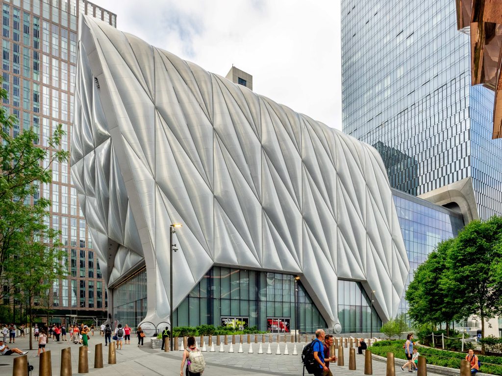 A modern building with a unique, silver, quilted exterior stands among high-rises. People walk and gather outside. The building features angular, geometric patterns and large glass windows. Trees and greenery are nearby.