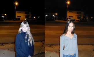 Two women stand side by side on a sidewalk at night, with a street and buildings in the background. The woman on the left wears a dark coat, and the woman on the right wears a gray top and jeans. Both face the camera.