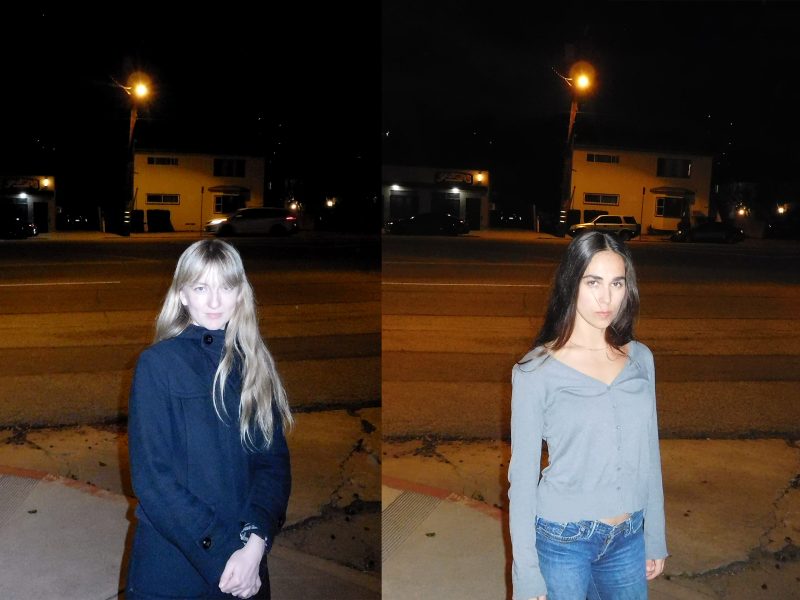 Two women stand side by side on a sidewalk at night, with a street and buildings in the background. The woman on the left wears a dark coat, and the woman on the right wears a gray top and jeans. Both face the camera.