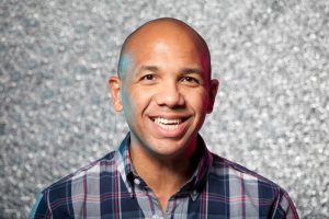 A smiling man with a shaved head wearing a blue plaid shirt stands in front of a sparkly, silver background at an AIGA Boston event featuring Rick Griffith at the ICA.