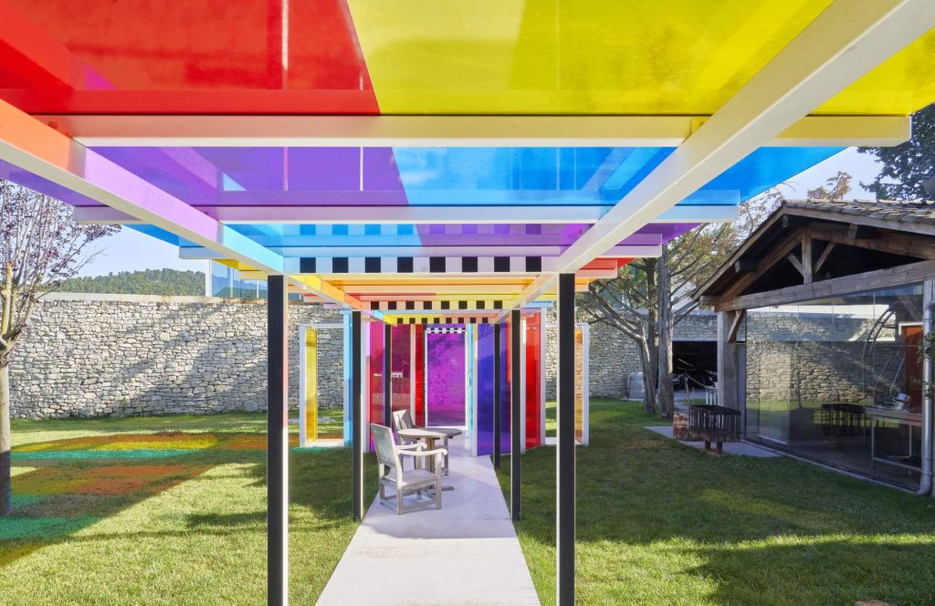 A colorful outdoor walkway features a roof made of translucent panels in red, yellow, blue, and purple, casting vibrant shadows on the ground. A chair and table sit beneath the structure in a grassy courtyard.