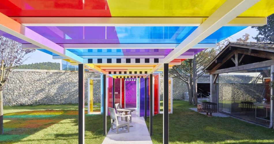 A colorful outdoor walkway features a roof made of translucent panels in red, yellow, blue, and purple, casting vibrant shadows on the ground. A chair and table sit beneath the structure in a grassy courtyard.