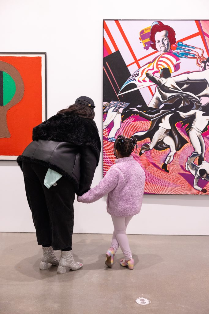 A woman and a young girl hold hands while viewing colorful artwork in a gallery. The woman wears a black jacket and silver boots; the girl is dressed in a pink outfit. They stand closely, looking at a painting.