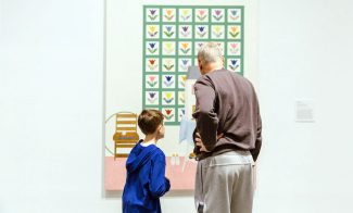 An older man and a young boy stand closely together, viewing a colorful painting with a grid of floral patterns in an art gallery. Their backs are to the camera as they observe the artwork.