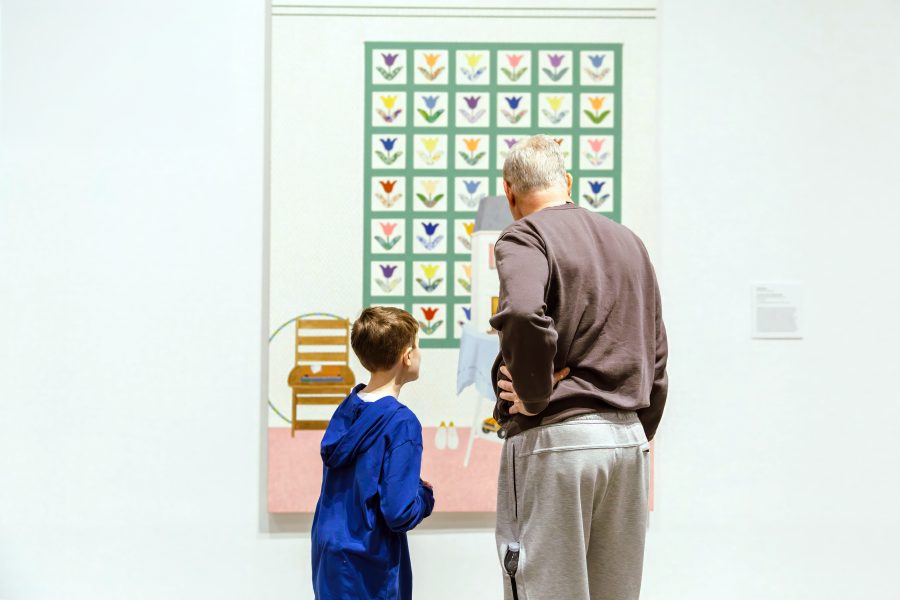 An older man and a young boy stand closely together, viewing a colorful painting with a grid of floral patterns in an art gallery. Their backs are to the camera as they observe the artwork.
