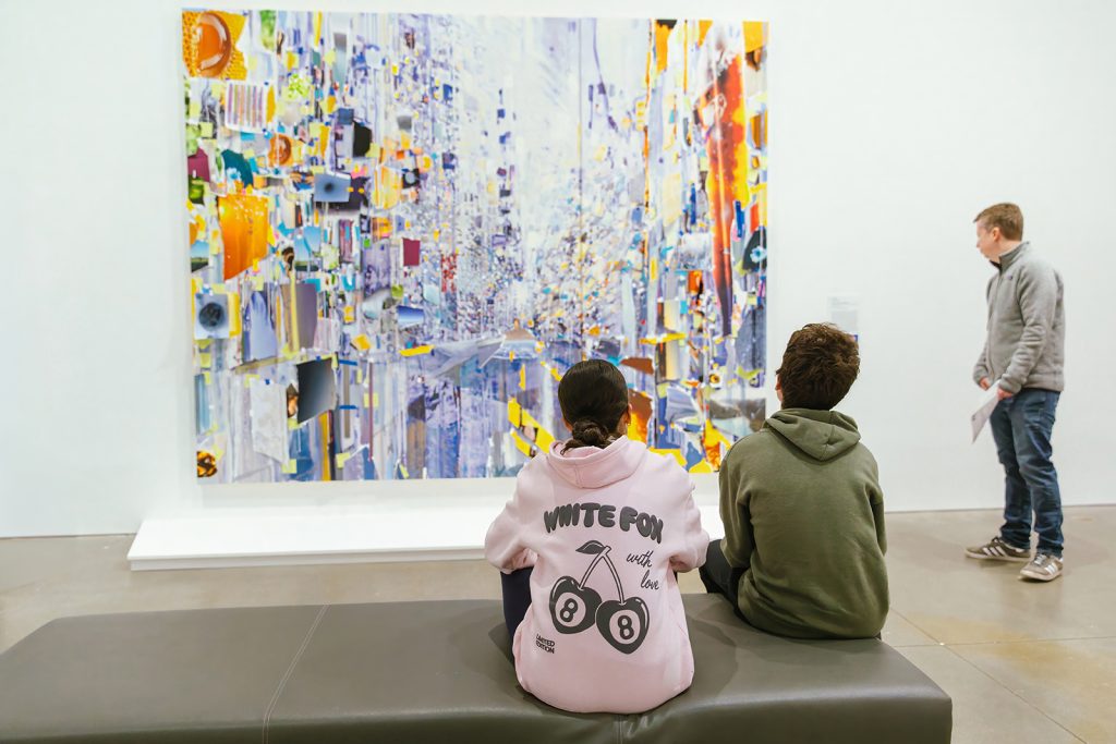 Two young people sit on a bench facing a large, colorful abstract painting, while another person stands to the side of it
