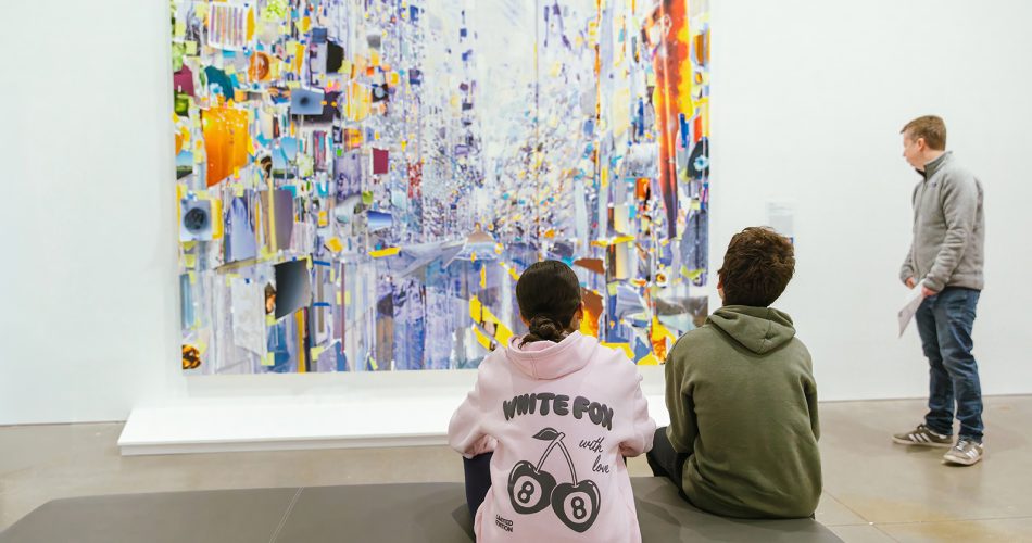Two young people sit on a bench facing a large, colorful abstract painting, while another person stands to the side of it