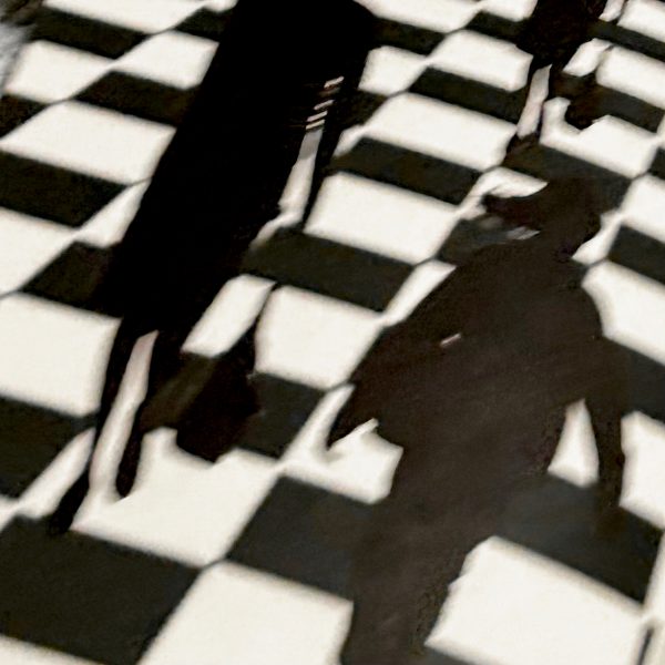 Silhouetted figures and their shadows are cast onto a black and white checkered floor, creating an abstract and dynamic scene.