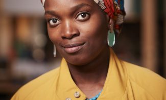 A headshot of Njideka Akunyili Crosby, wearing a colorful patterned headwrap, turquoise earrings, a blue necklace, a yellow coat, and a magenta top. She has a slight smile and is standing indoors with a blurred background.
