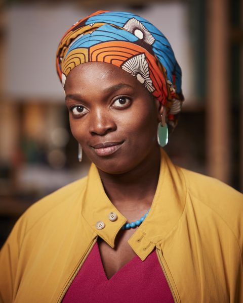 A headshot of Njideka Akunyili Crosby, wearing a colorful patterned headwrap, turquoise earrings, a blue necklace, a yellow coat, and a magenta top. She has a slight smile and is standing indoors with a blurred background.