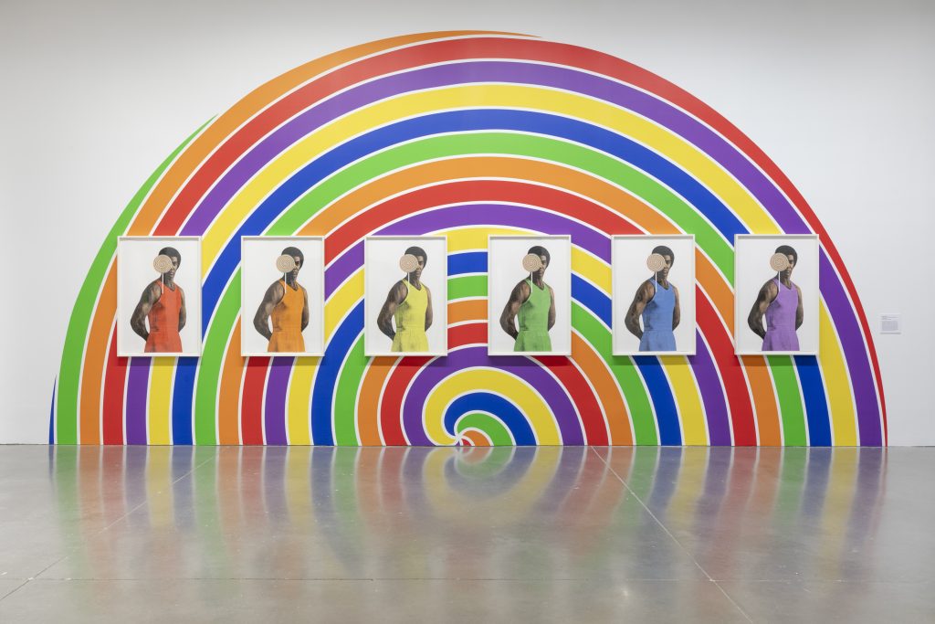 Five portraits of the same person, each in a different color outfit, are displayed on a wall painted with a vibrant rainbow spiral