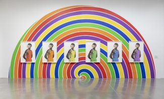 Five portraits of the same person, each in a different color outfit, are displayed on a wall painted with a vibrant rainbow spiral