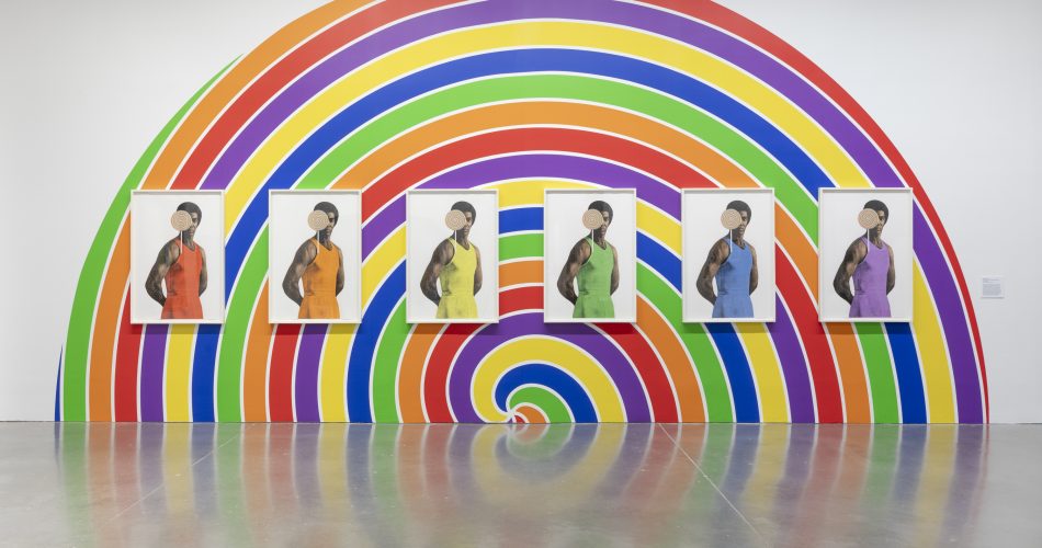Five portraits of the same person, each in a different color outfit, are displayed on a wall painted with a vibrant rainbow spiral