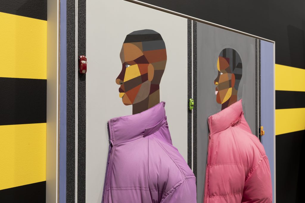 A side view of a paintiing featuring two side-by-side profiles of two women with geometric, multicolored facial features, each wearing a real puffer jacket—one purple and one pink—protruding from the canvas. The wall it hangs on has black and yellow stripes.