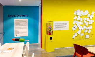 A brightly colored room with a teal wall labeled Extra/Ordinary, a yellow wall with papers pinned on a grid, a gumball machine, and playful furniture. The modern space is part of the Extra/Ordinary ICA Boston Mediatheque.