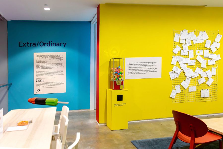 A brightly colored room with a teal wall labeled Extra/Ordinary, a yellow wall with papers pinned on a grid, a gumball machine, and playful furniture. The modern space is part of the Extra/Ordinary ICA Boston Mediatheque.