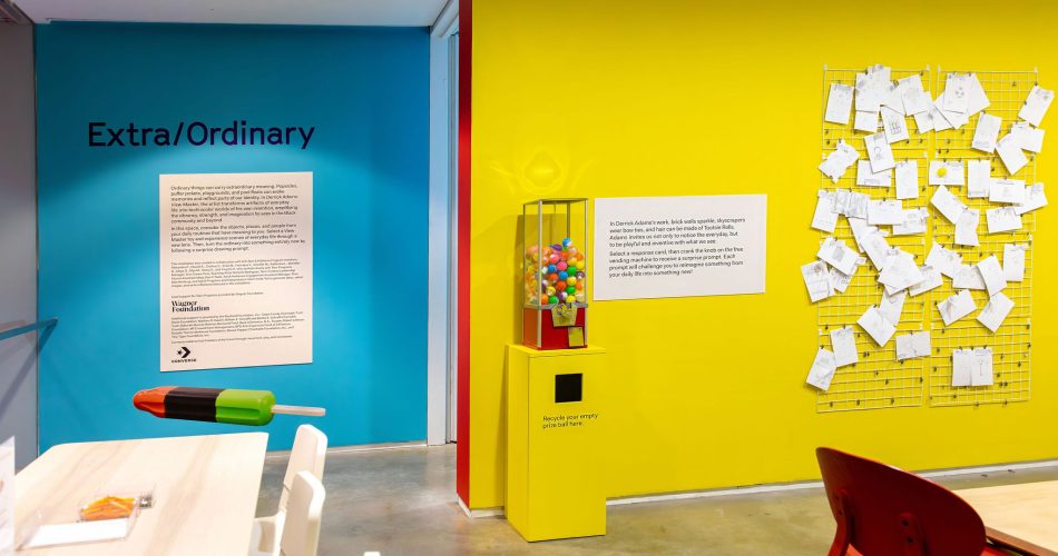 A brightly colored room with a teal wall labeled Extra/Ordinary, a yellow wall with papers pinned on a grid, a gumball machine, and playful furniture. The modern space is part of the Extra/Ordinary ICA Boston Mediatheque.