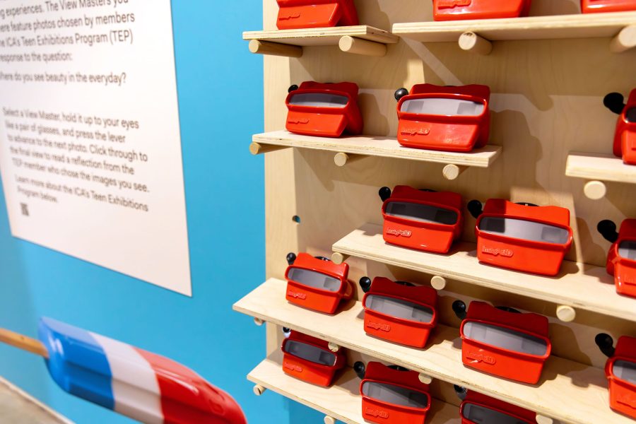 Rows of red plastic viewfinders are displayed on light wooden shelves at the Extra/Ordinary ICA Boston Mediatheque, with a blue section and informational text visible to the side.