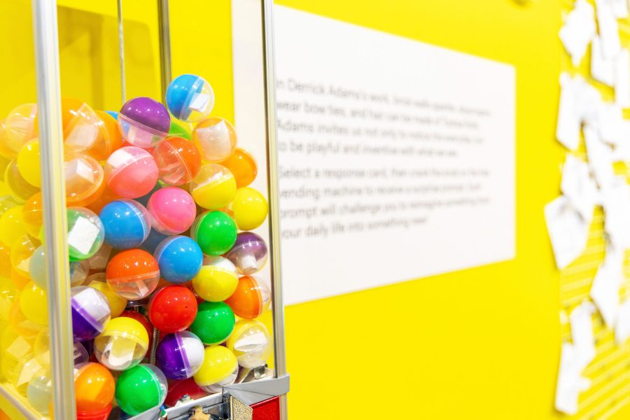 A close-up of a vending machine filled with colorful plastic capsules against a bright yellow wall at the Extra/Ordinary ICA Boston Mediatheque, with text and papers blurred in the background.