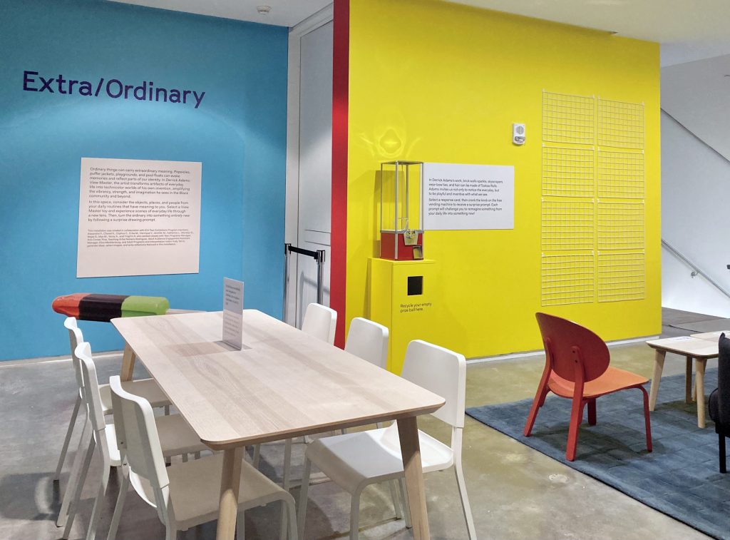 A room with blue and yellow painted walls with text reading Extra/Ordinary, text displays, and a long wooden table with white chairs