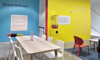 A room with blue and yellow painted walls with text reading Extra/Ordinary, text displays, and a long wooden table with white chairs