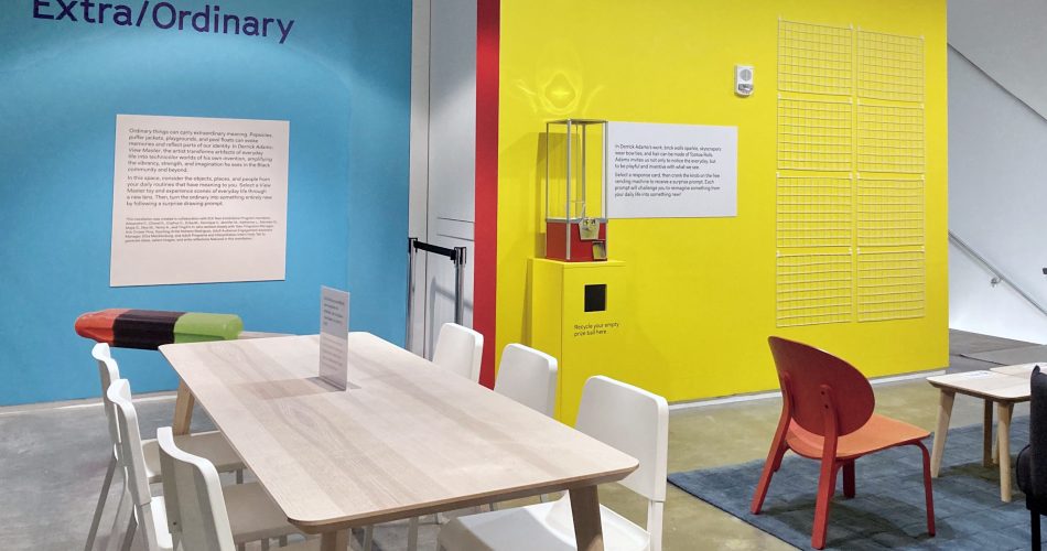 A room with blue and yellow painted walls with text reading Extra/Ordinary, text displays, and a long wooden table with white chairs
