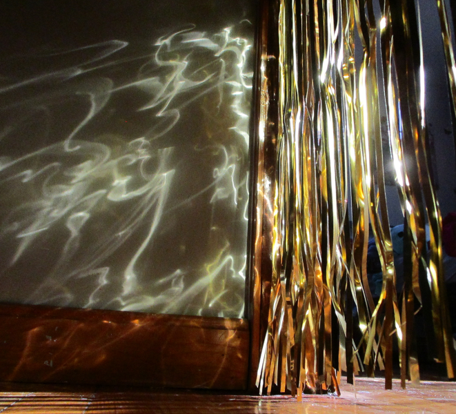 Golden metallic streamers hang beside a wooden-framed surface, casting wavy, shimmering patterns onto a pale wall and floor