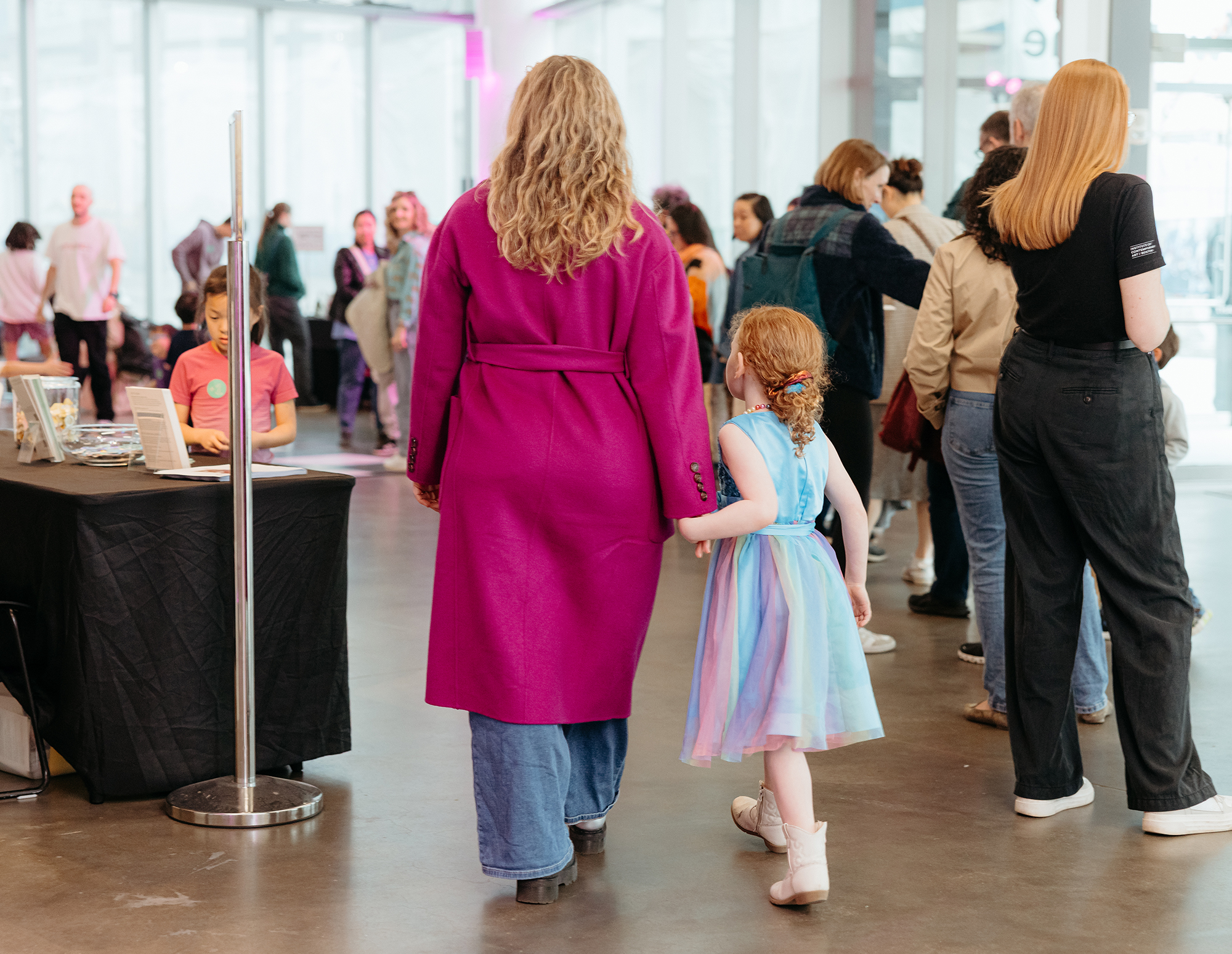 A woman in a bright magenta coat holds hands with a young girl in a colorful dress as they walk through the busy ICA lobby, with other people and display tables visible around them.