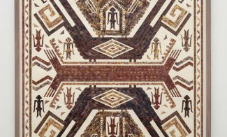 A framed artwork features a symmetrical, geometric pattern with bird-like and humanoid shapes in earthy tones of brown, red, and beige, resembling traditional or folk textile designs on a white background.
