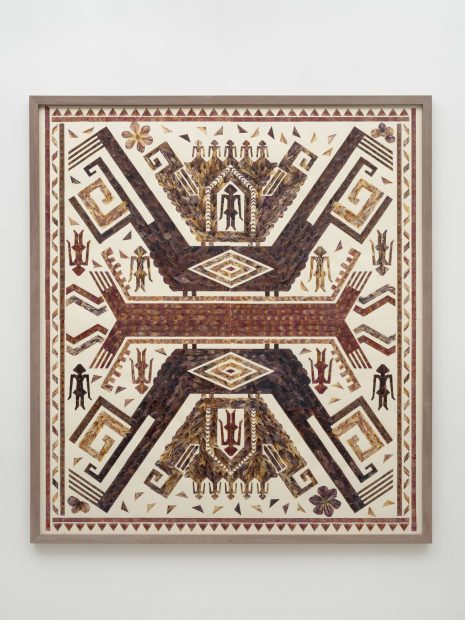 A framed artwork features a symmetrical, geometric pattern with bird-like and humanoid shapes in earthy tones of brown, red, and beige, resembling traditional or folk textile designs on a white background.