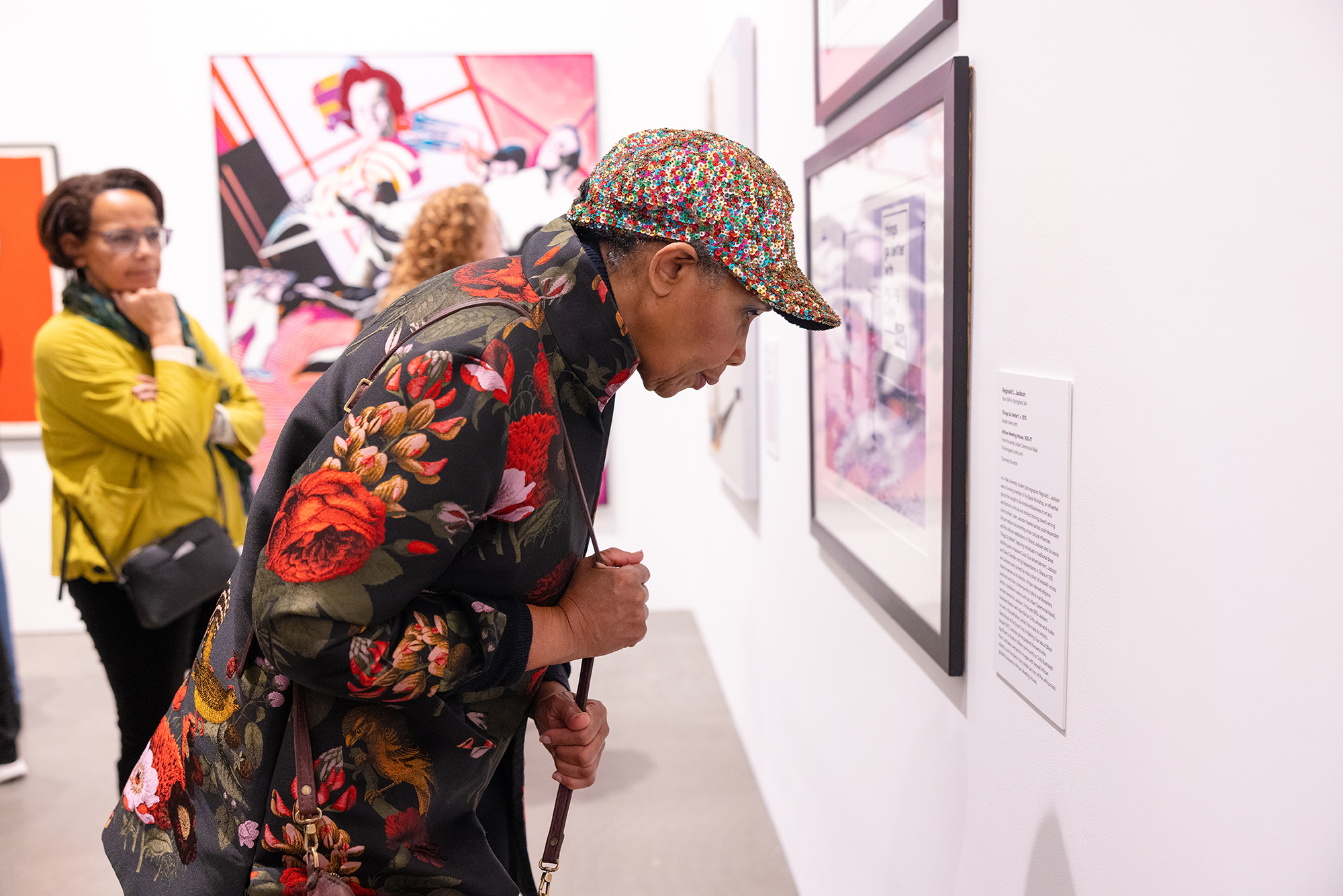 A woman in a floral jacket and patterned cap leans in to closely examine framed artwork on a gallery wall, while two other visitors observe the displays in the background.