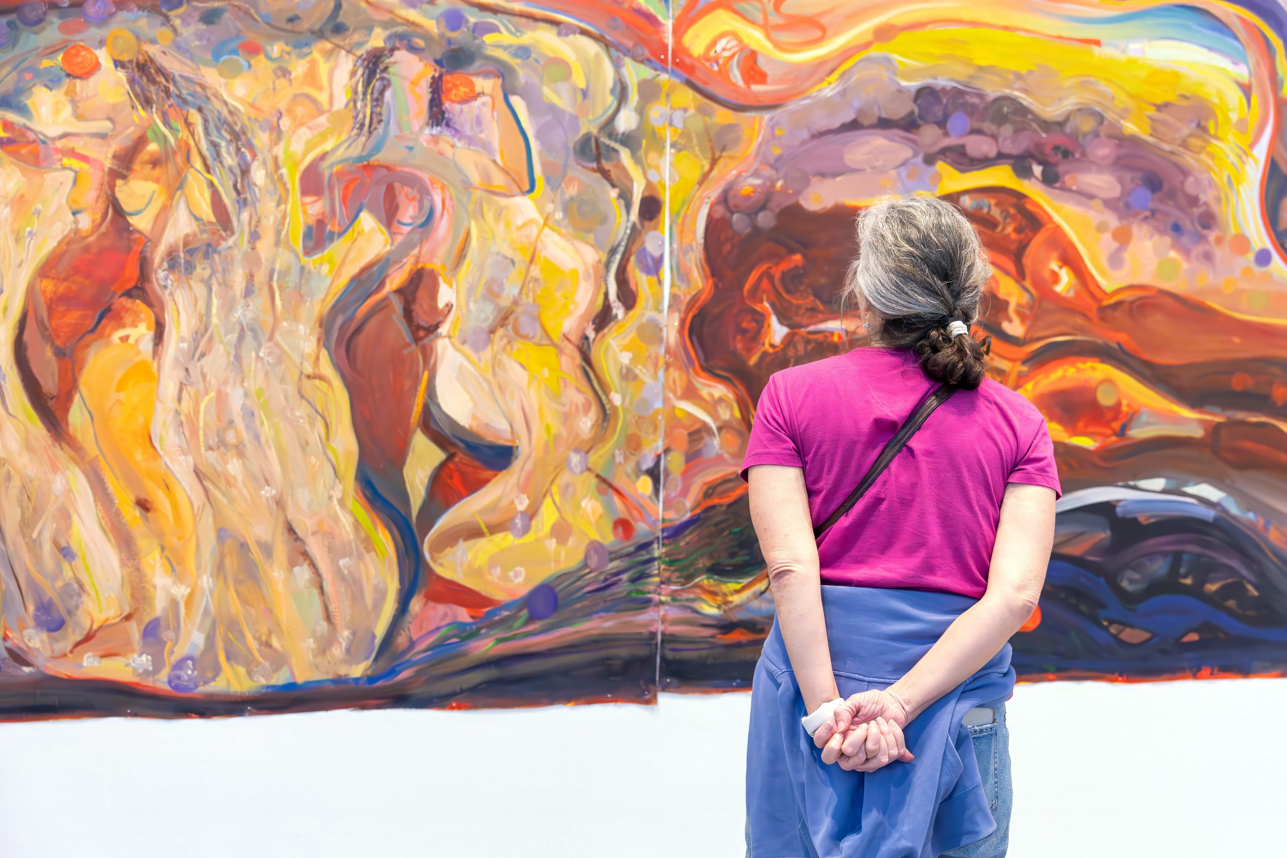 A woman with gray hair in a ponytail, wearing a pink shirt and blue sweater tied around her waist, stands with hands clasped behind her back, admiring a large, colorful abstract painting
