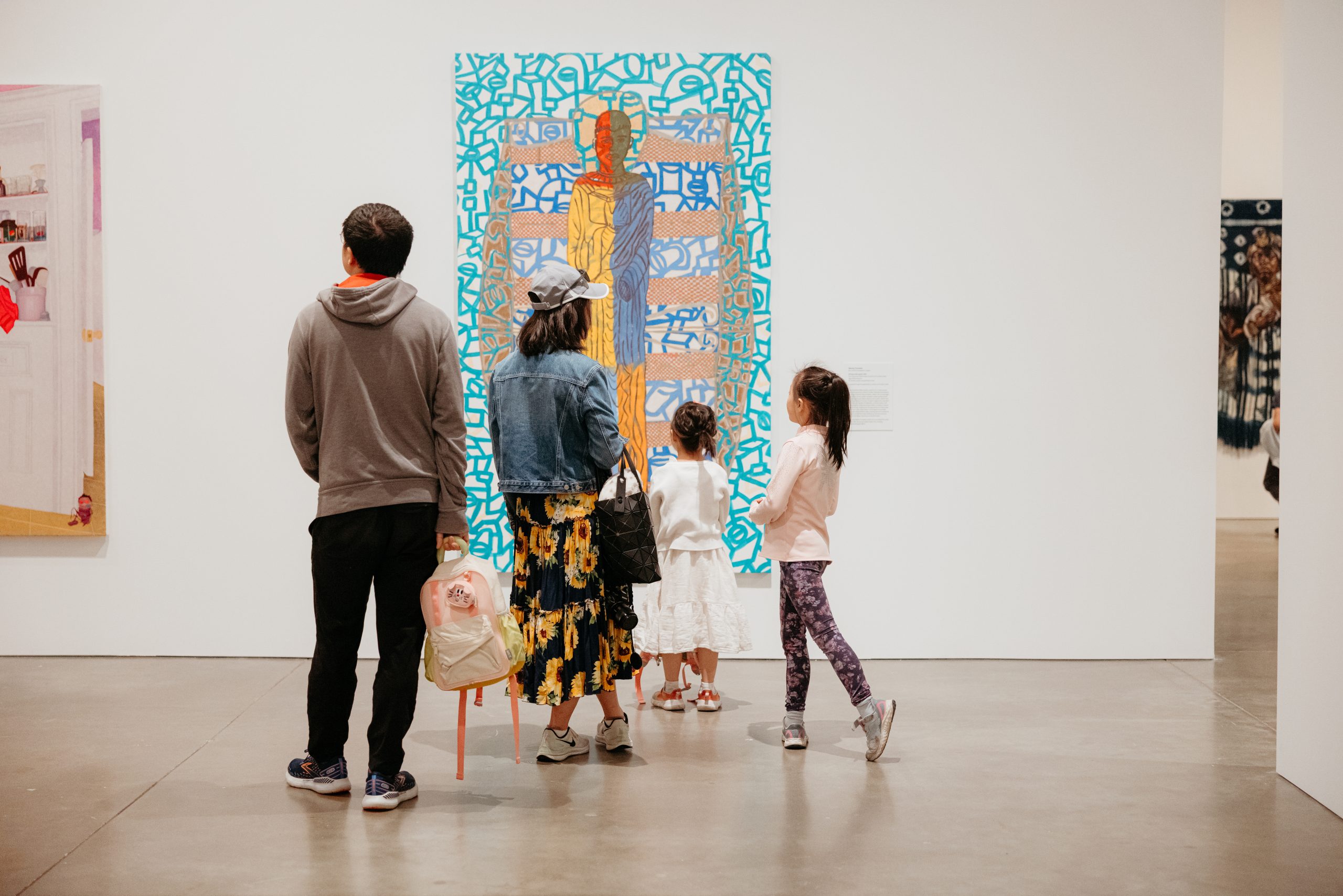 Four people, including two children, stand in an art gallery looking at a colorful painting on a white wall