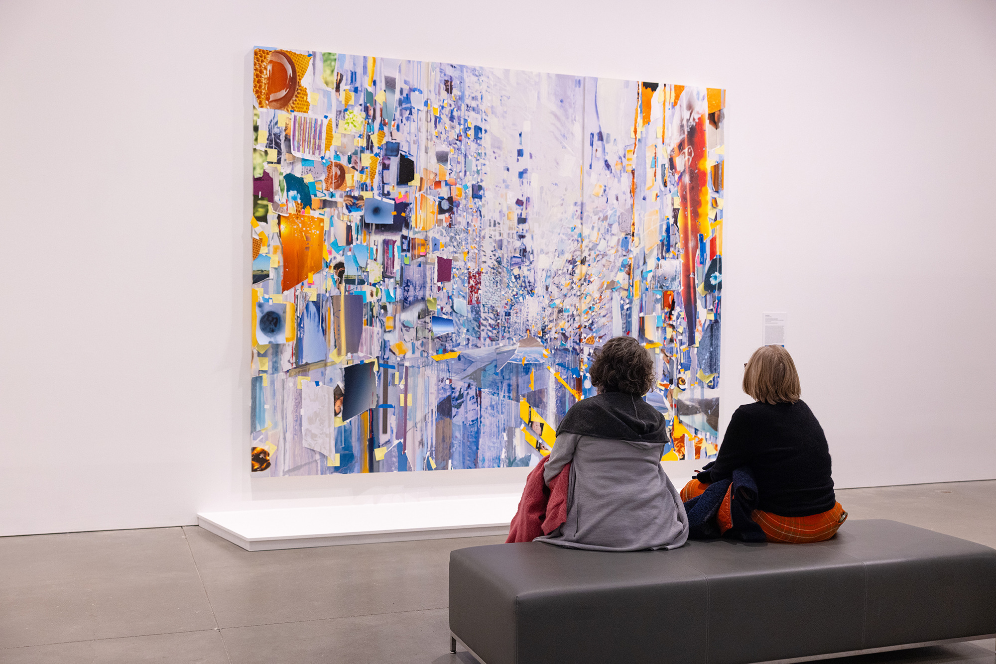 Two people sit on a bench in a gallery, viewing a large, colorful abstract painting with vibrant splashes of blue, orange, and yellow on a white wall