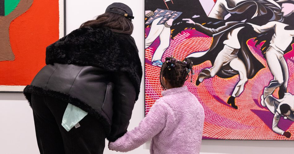An adult in a black jacket and cap holds hands with a child in a lavender jacket as they admire colorful art in the gallery.