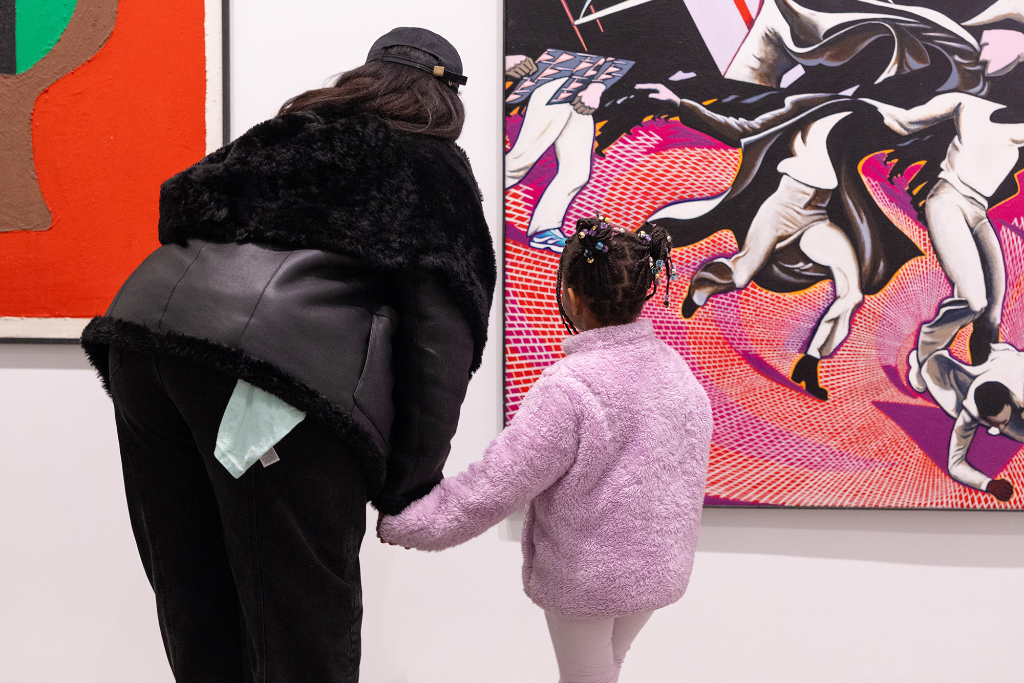 An adult in a black jacket and cap holds hands with a child in a lavender jacket as they admire colorful art in the gallery.