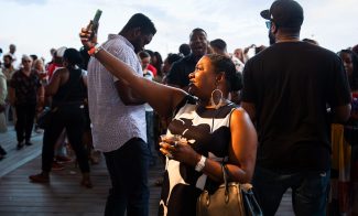 A woman in a black and white dress takes a selfie with her phone at an outdoor event, surrounded by a crowd of people on a wooden deck during daylight.