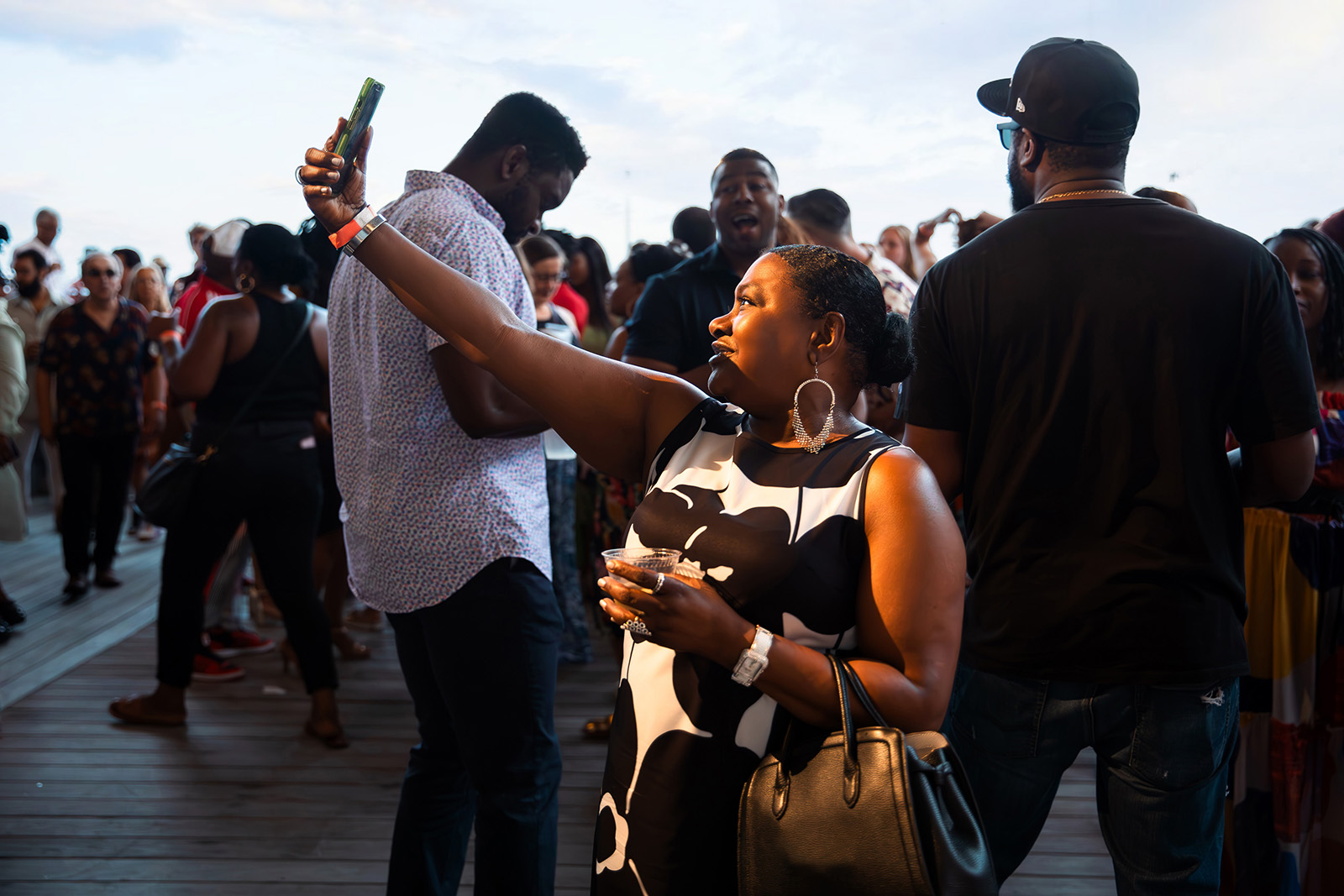 A woman in a black and white dress takes a selfie with her phone at an outdoor event, surrounded by a crowd of people on a wooden deck during daylight.