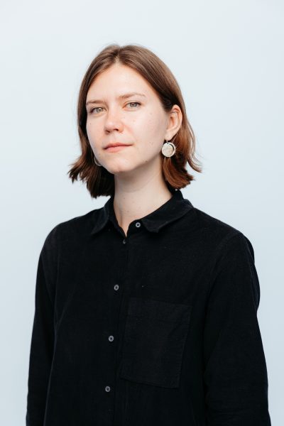 A woman with short brown hair, wearing a black button-up shirt and large round earrings, stands against a plain light background, looking calmly at the camera with a slight smile.
