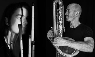 Black and white photo of a woman with woodwind instruments casting shadows on her face, and a man holding and looking at a large wind instrument against a dark background.