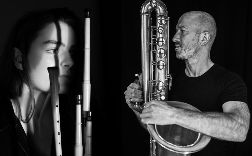 Black and white photo of a woman with woodwind instruments casting shadows on her face, and a man holding and looking at a large wind instrument against a dark background.