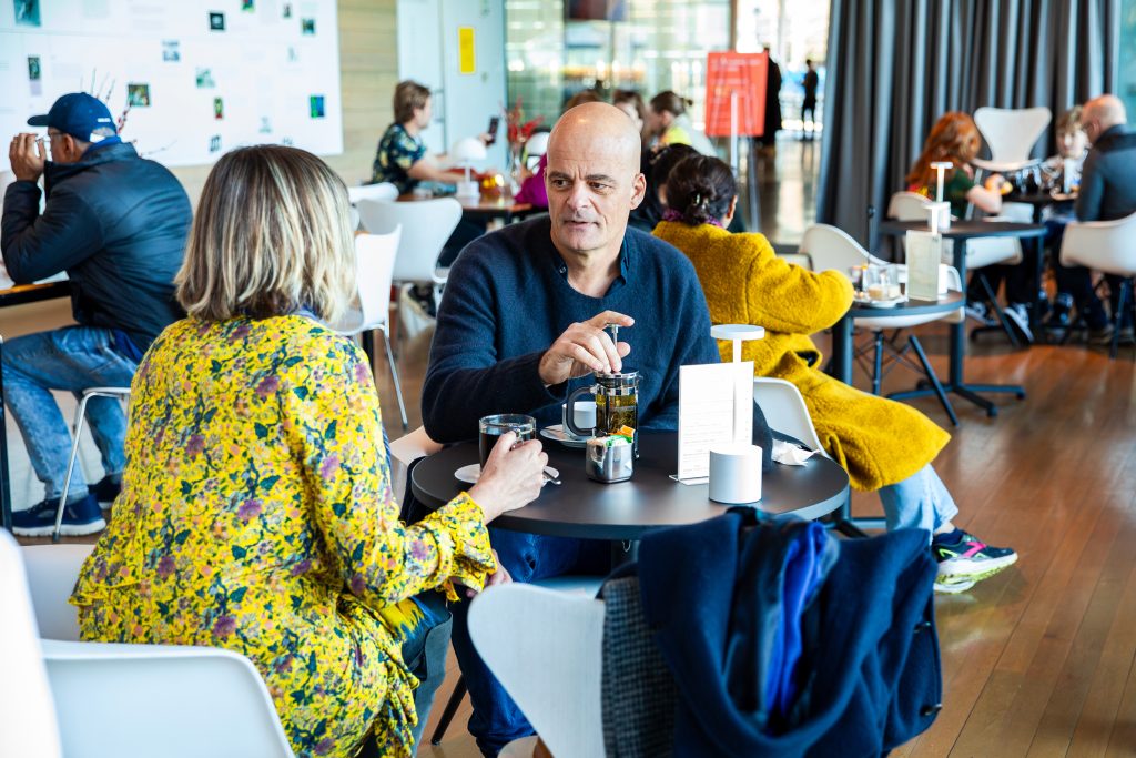A man and a woman sit at a round table in a busy, modern café at the Institute of Contemporary Art Boston, talking over drinks. The woman wears a yellow floral dress; the man has a dark sweater. Other patrons chat in the background.