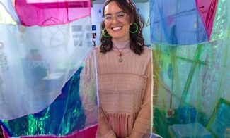 A person with glasses, large green hoop earrings, and a light pink dress smiles while standing between colorful, translucent fabric panels in a bright, creative indoor space.