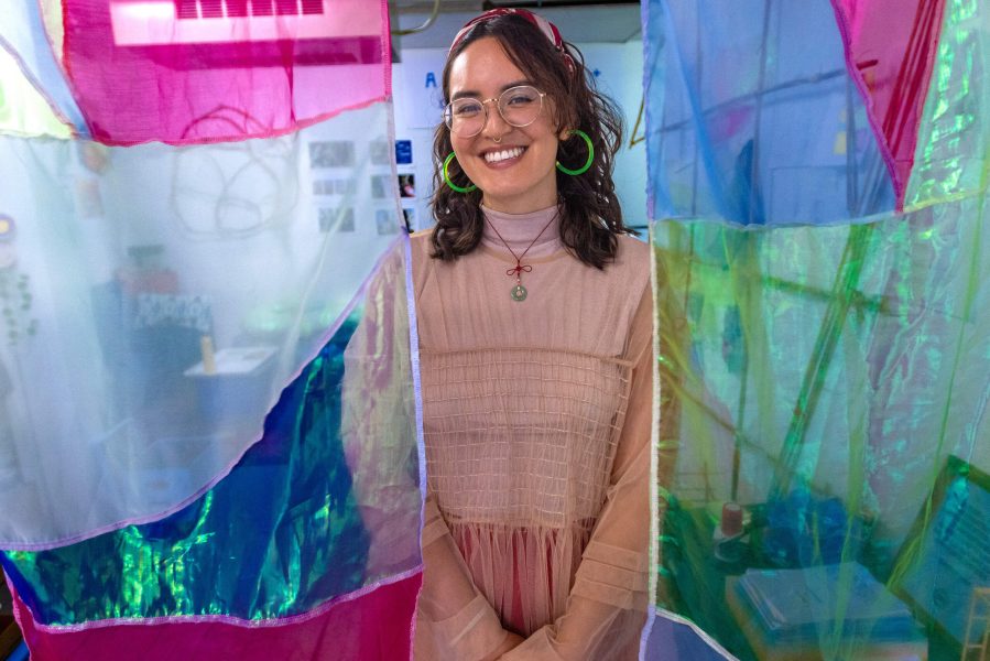 A person with glasses, large green hoop earrings, and a light pink dress smiles while standing between colorful, translucent fabric panels in a bright, creative indoor space.
