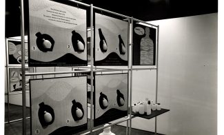 Black and white photo of an exhibit with illustrated panels showing sequential diagrams of a heart and blood flow. Nearby, a table holds several plastic bottles of varying sizes. The display is set in a gallery space.