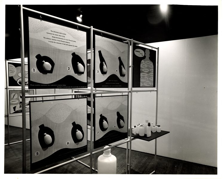 Black and white photo of an exhibit with illustrated panels showing sequential diagrams of a heart and blood flow. Nearby, a table holds several plastic bottles of varying sizes. The display is set in a gallery space.