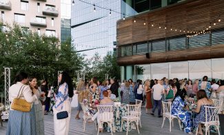A large group of people dressed in summer attire gather on a modern outdoor terrace, socializing and sitting at tables under string lights, with tall buildings and greenery in the background.